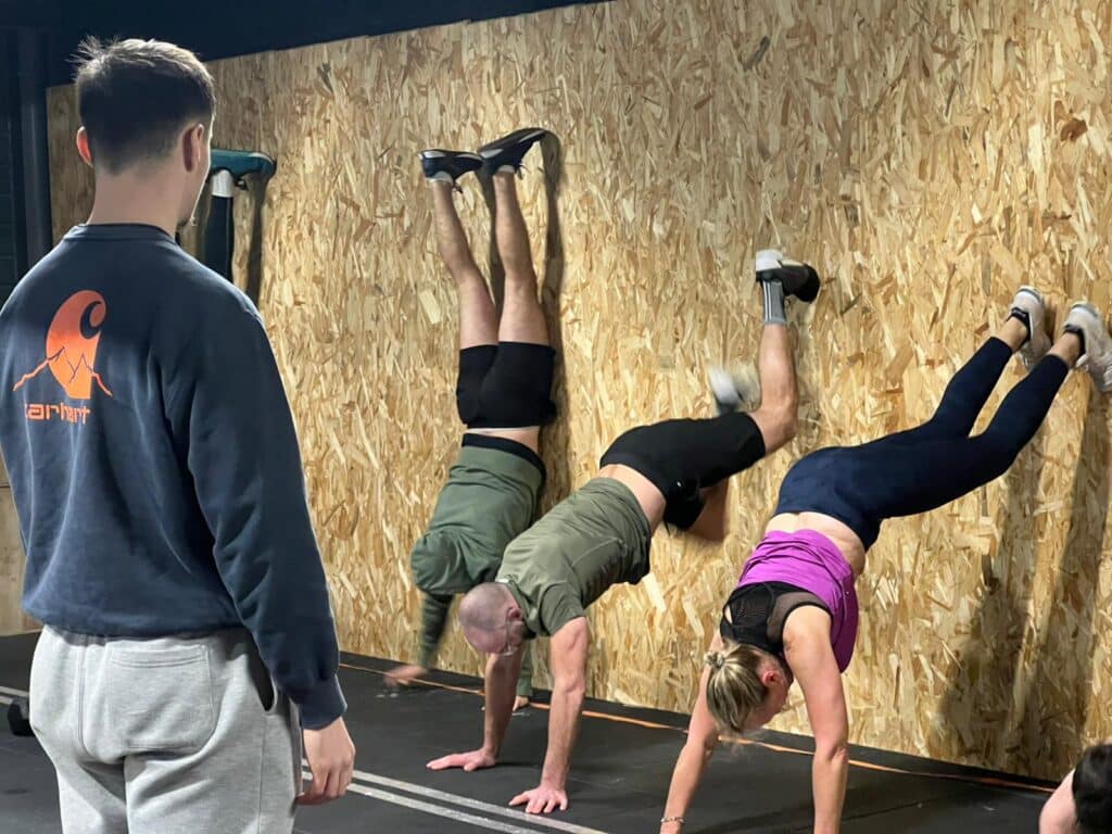 Groupe de personnes font des poiriers contre un mur en bois pendant une séance de fitness, un coach observe.