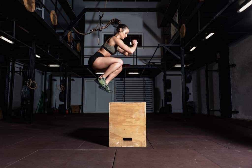 Saut sur boîte Cross Training : Entraînement intense pour femme. Athlète femme effectuant un saut sur boîte en suspension dans une salle de musculation sombre.