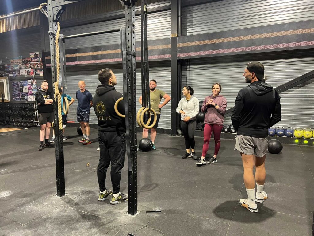 CrossFit : Entraînement de Groupe Dirigé par Coach Groupe d'athlètes écoutant un coach dans une salle de CrossFit avec rig et rings. Texte visible : HLTΣ.