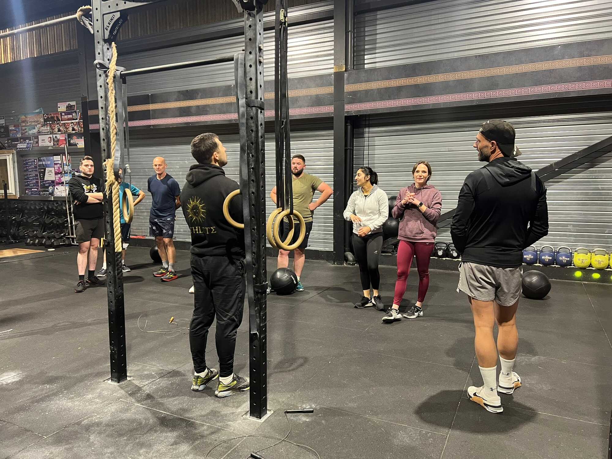 Groupe d'athlètes écoutant un coach dans une salle de CrossFit avec rig et rings. Texte visible : HLTΣ.
