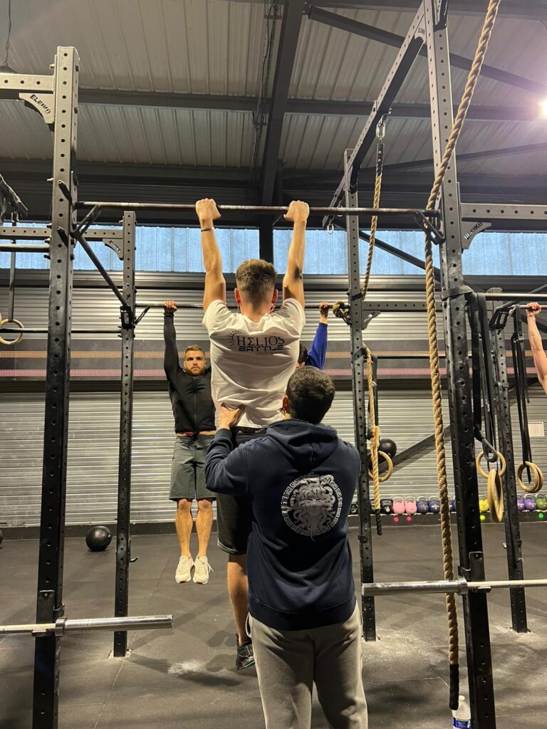 Gym: athlète en t-shirt Helios Battle fait une traction assistée par un partenaire. D'autres s'entraînent aux tractions sur la barre.