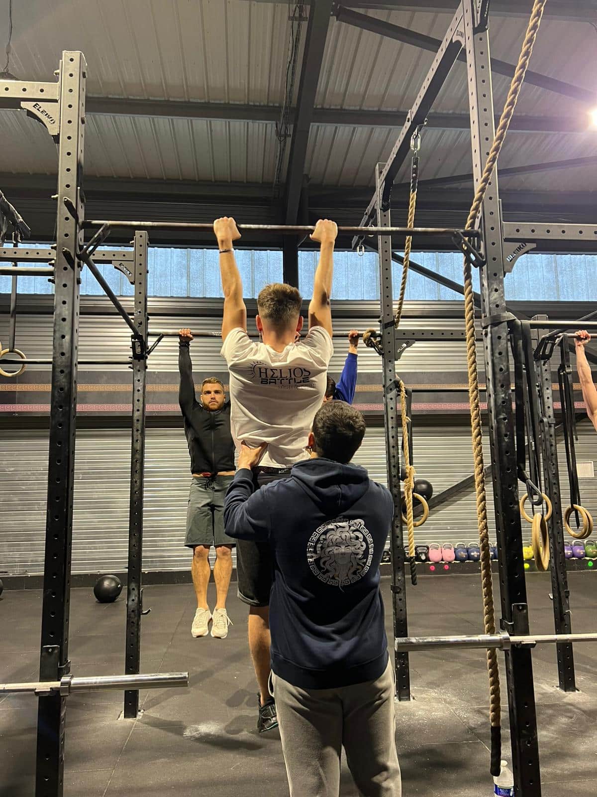 Gym: athlète en t-shirt Helios Battle fait une traction assistée par un partenaire. D'autres s'entraînent aux tractions sur la barre.